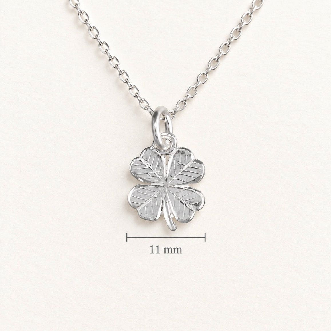 A silver four-leaf clover pendant necklace on a matching chain, shown against a light off-white background with a '11 mm' size indicator line and text below the pendant.