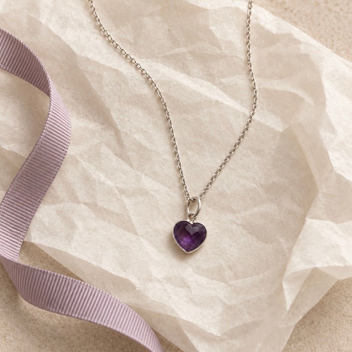 A silver chain necklace with a heart-shaped purple amethyst gemstone pendant, styled on a textured beige surface with a lavender ribbon.