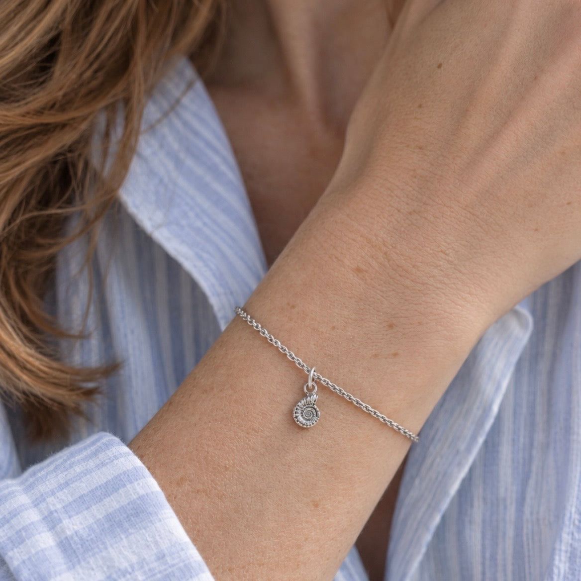 A close-up of a delicate silver chain bracelet with a small circular sunburst charm, worn on a woman's wrist. The person is wearing a light blue and white striped button-down shirt.