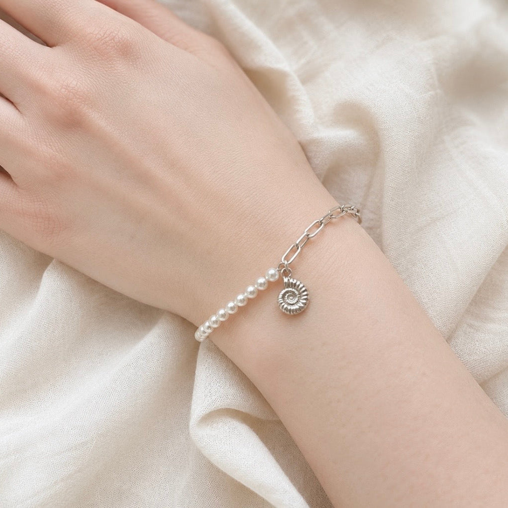A delicate silver paperclip chain bracelet with a strand of small white pearls and an ammonite fossil charm, shown worn on a person's wrist against a soft cream-colored fabric background.