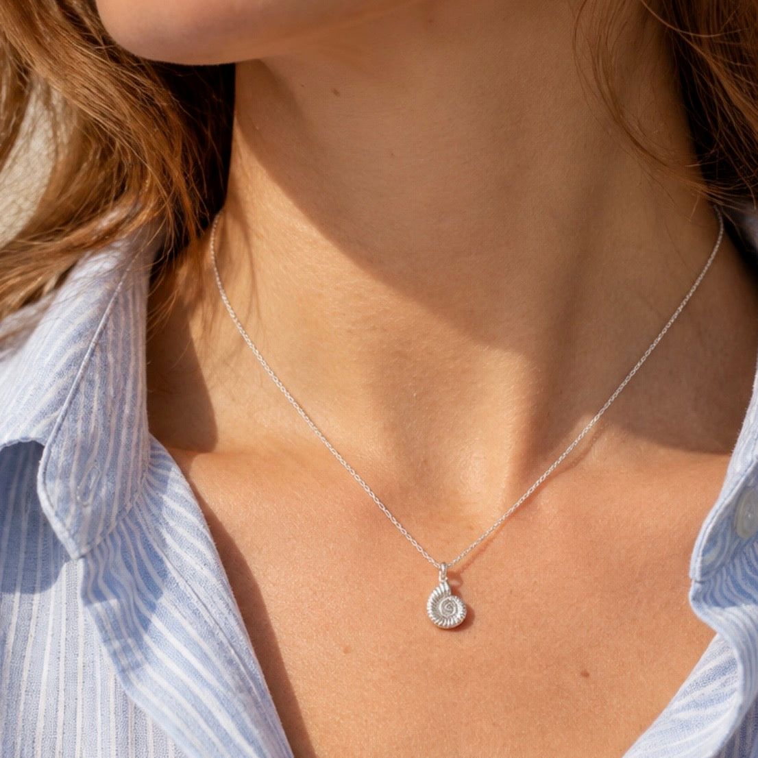 A close-up of a model wearing a delicate silver chain necklace with a spiral shell pendant, styled with a light blue and white striped shirt.