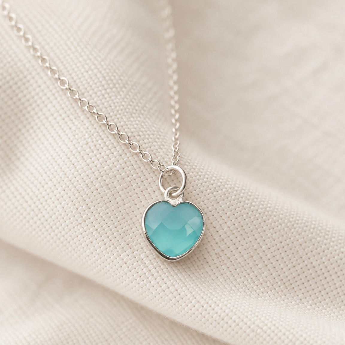 A silver heart pendant necklace with a faceted light blue Aqua Chalcedony gemstone, resting on a textured beige fabric background.