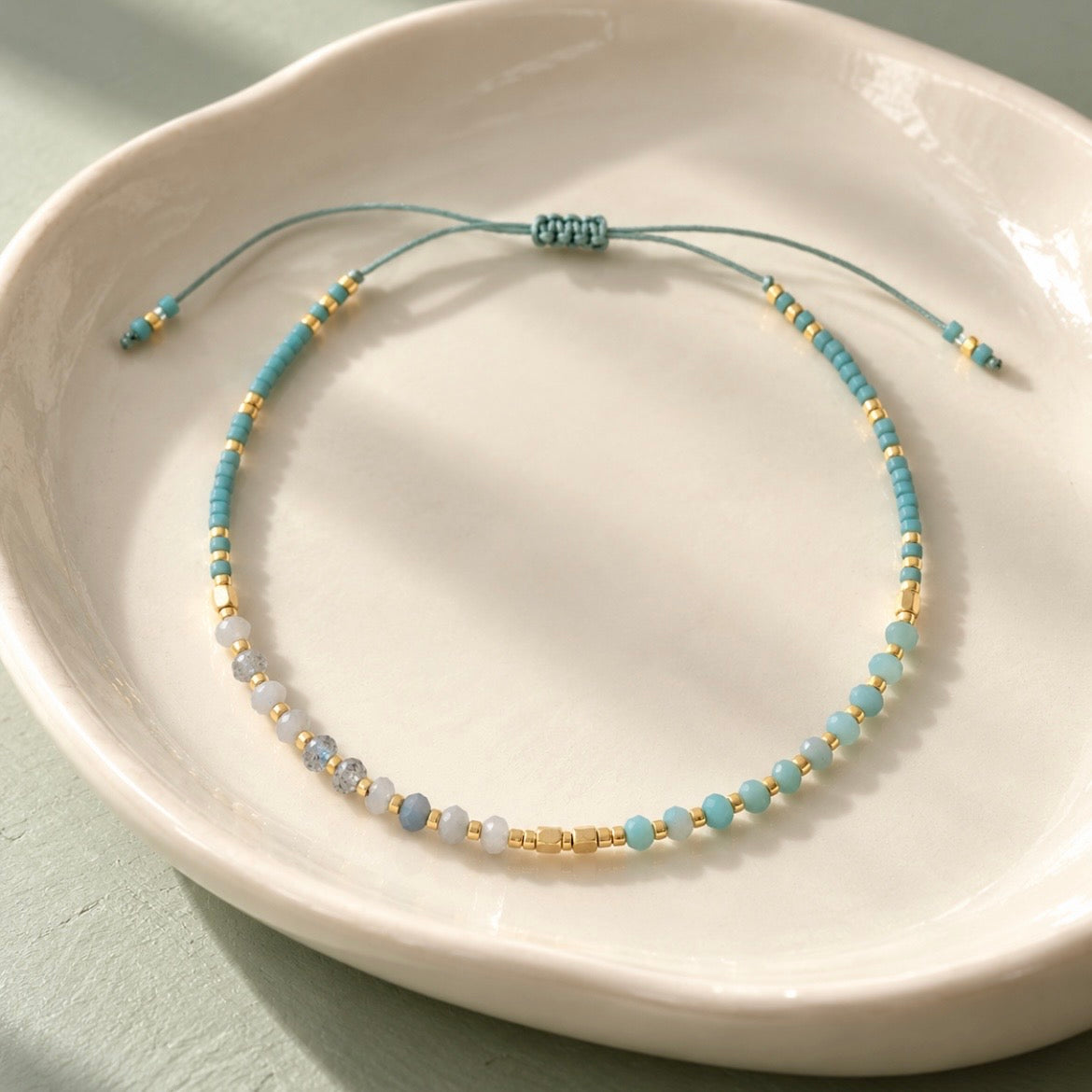 A delicate beaded cord bracelet with light blue beads, white beads, and small gold spacer beads, displayed on a white ceramic dish.