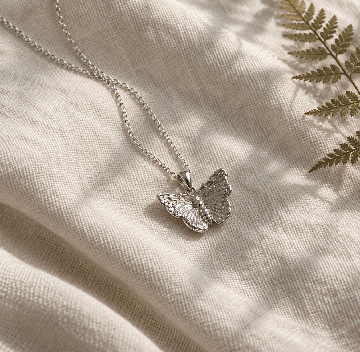 A silver butterfly pendant necklace on a delicate silver chain, displayed on a textured cream linen fabric with soft leaf shadows and a dried fern leaf.