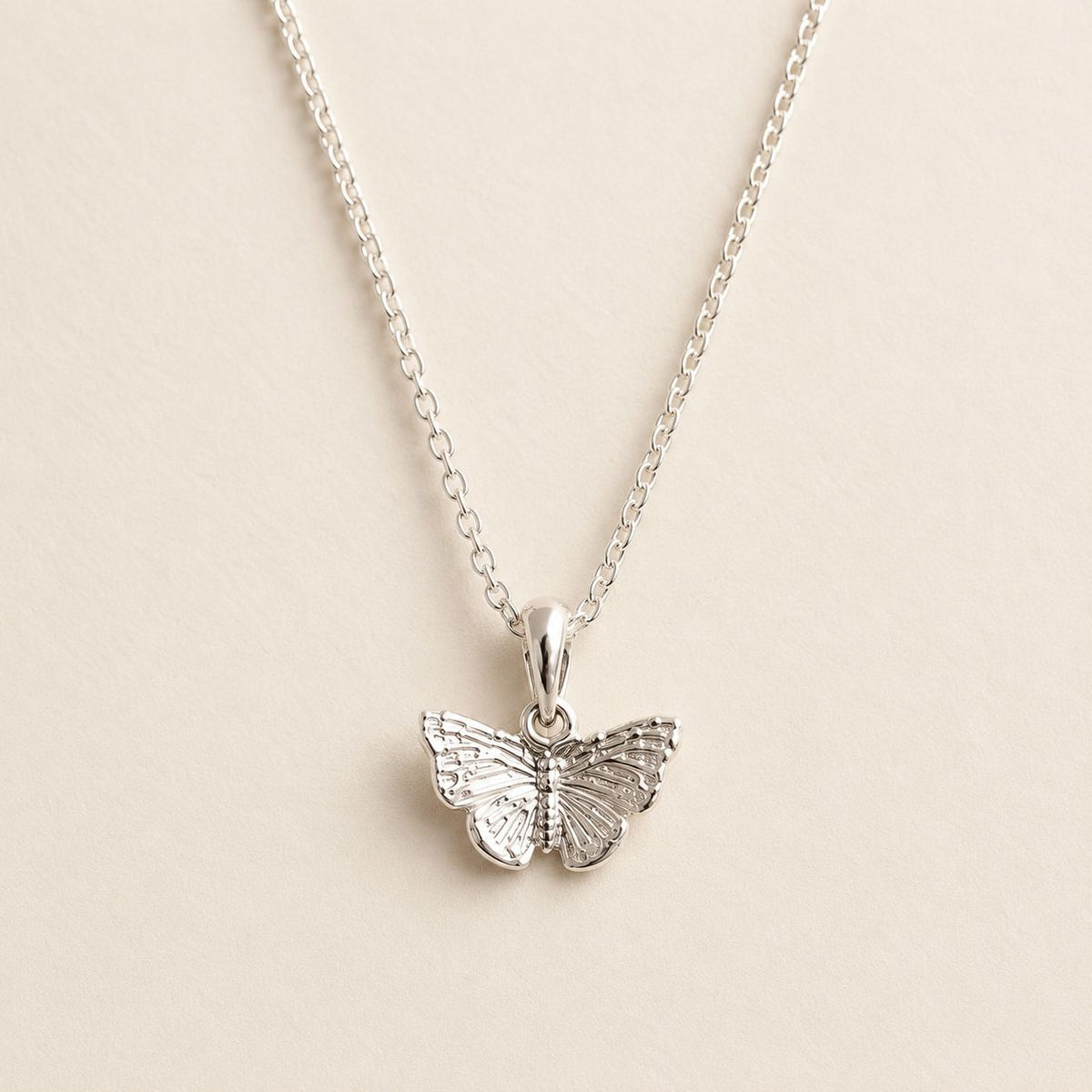 A silver butterfly pendant necklace on a delicate silver chain, displayed against a light beige textured background.