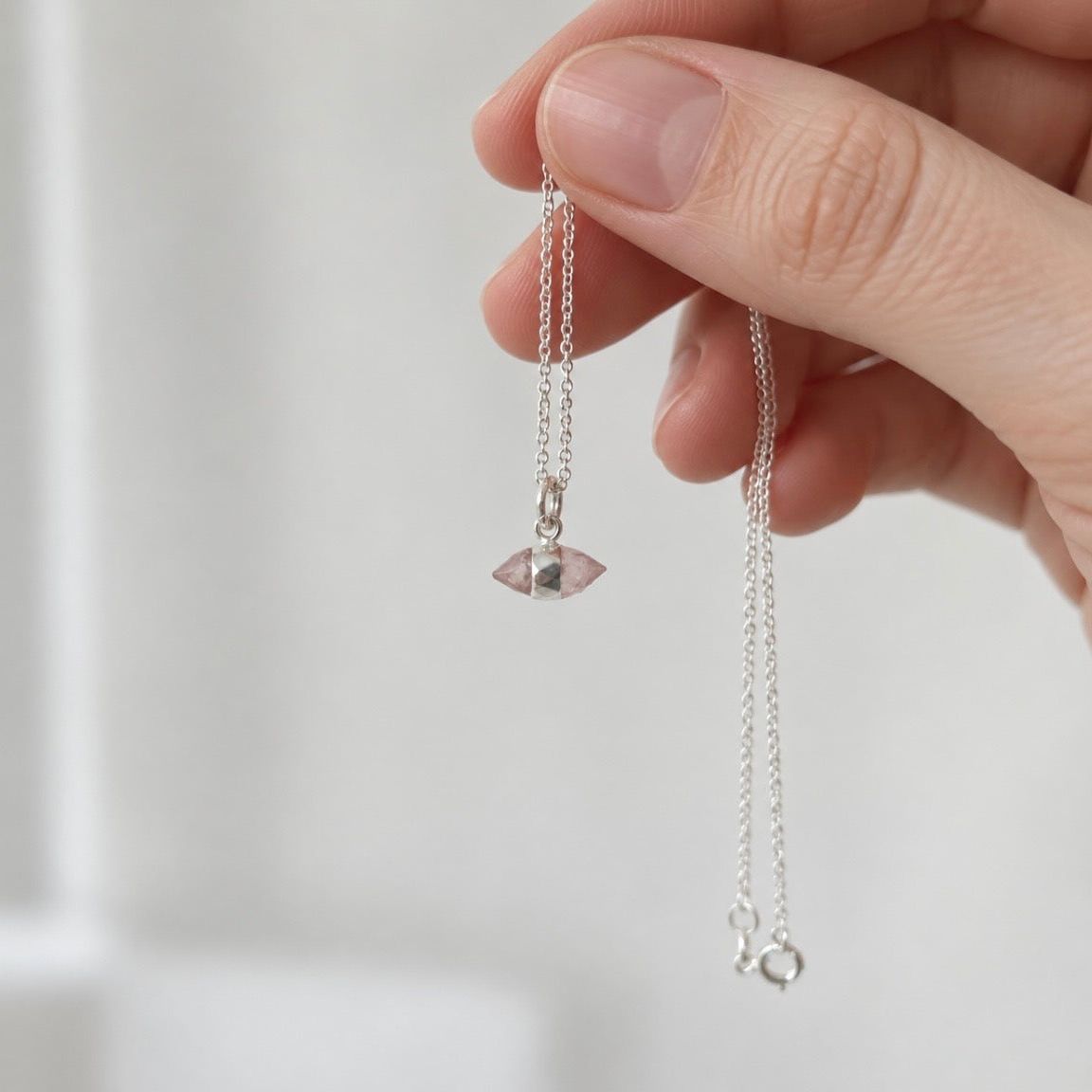 A close-up of a hand holding a delicate silver chain necklace with a small faceted pink rose quartz pendant against a soft, light gray background.
