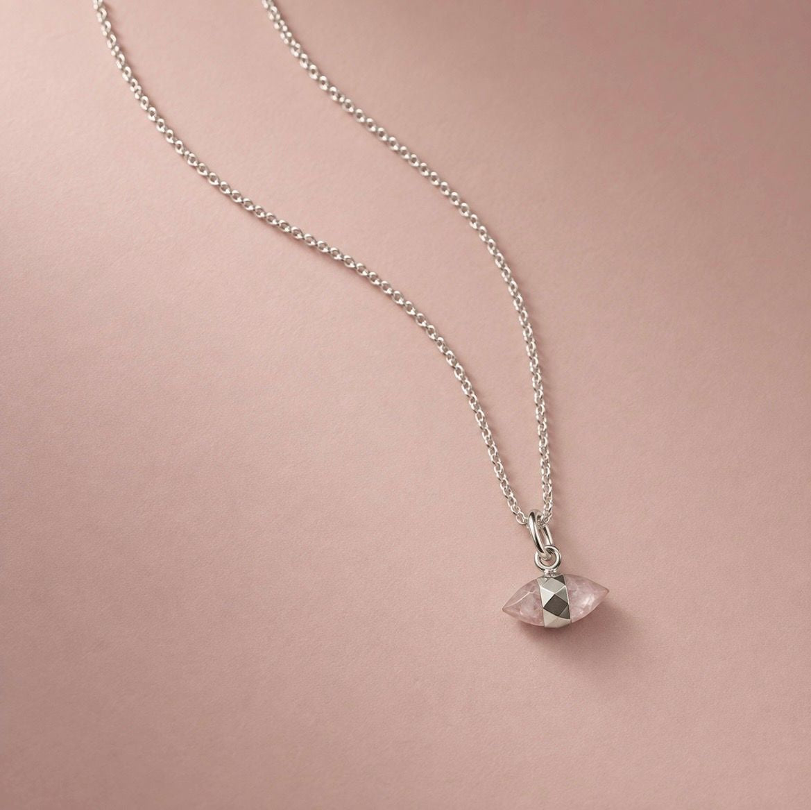 A silver chain necklace with a small faceted pink rose quartz pendant, displayed on a soft pink background.