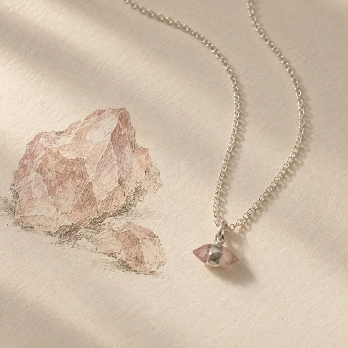 A silver chain necklace with a small faceted pink rose quartz crystal pendant, styled on a textured cream paper background next to a sketch of pink rocks.