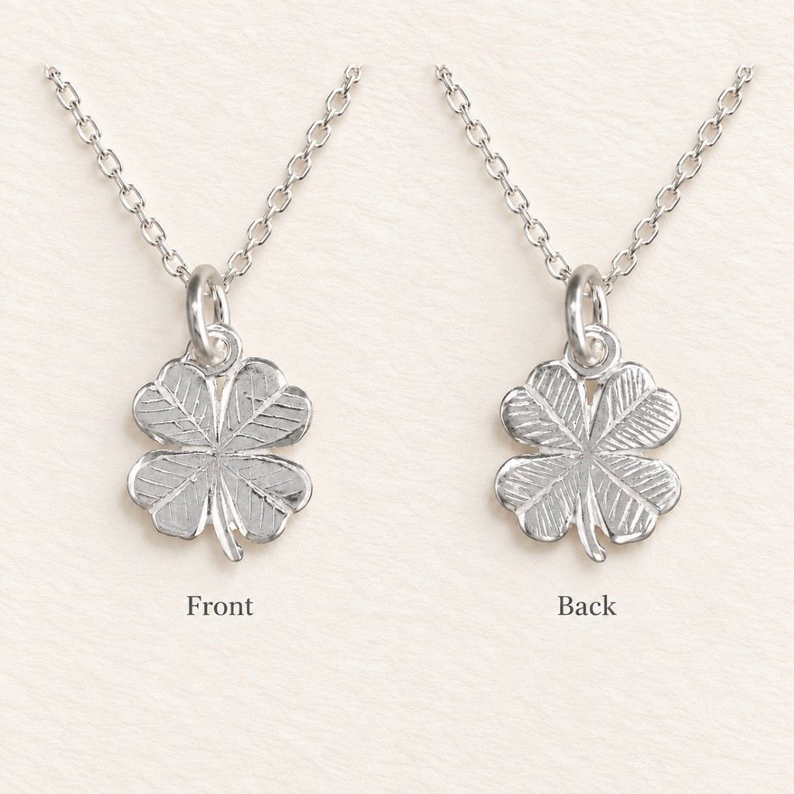 A silver four-leaf clover pendant necklace shown from both front and back on a light cream textured background. The image displays the polished front face on the left and the textured reverse side on the right, both hanging from a delicate silver link chain.