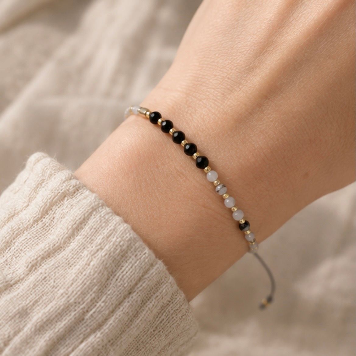 A delicate beaded bracelet with black, white, and gold beads worn on a person's wrist, shown against a soft beige knit sweater background.