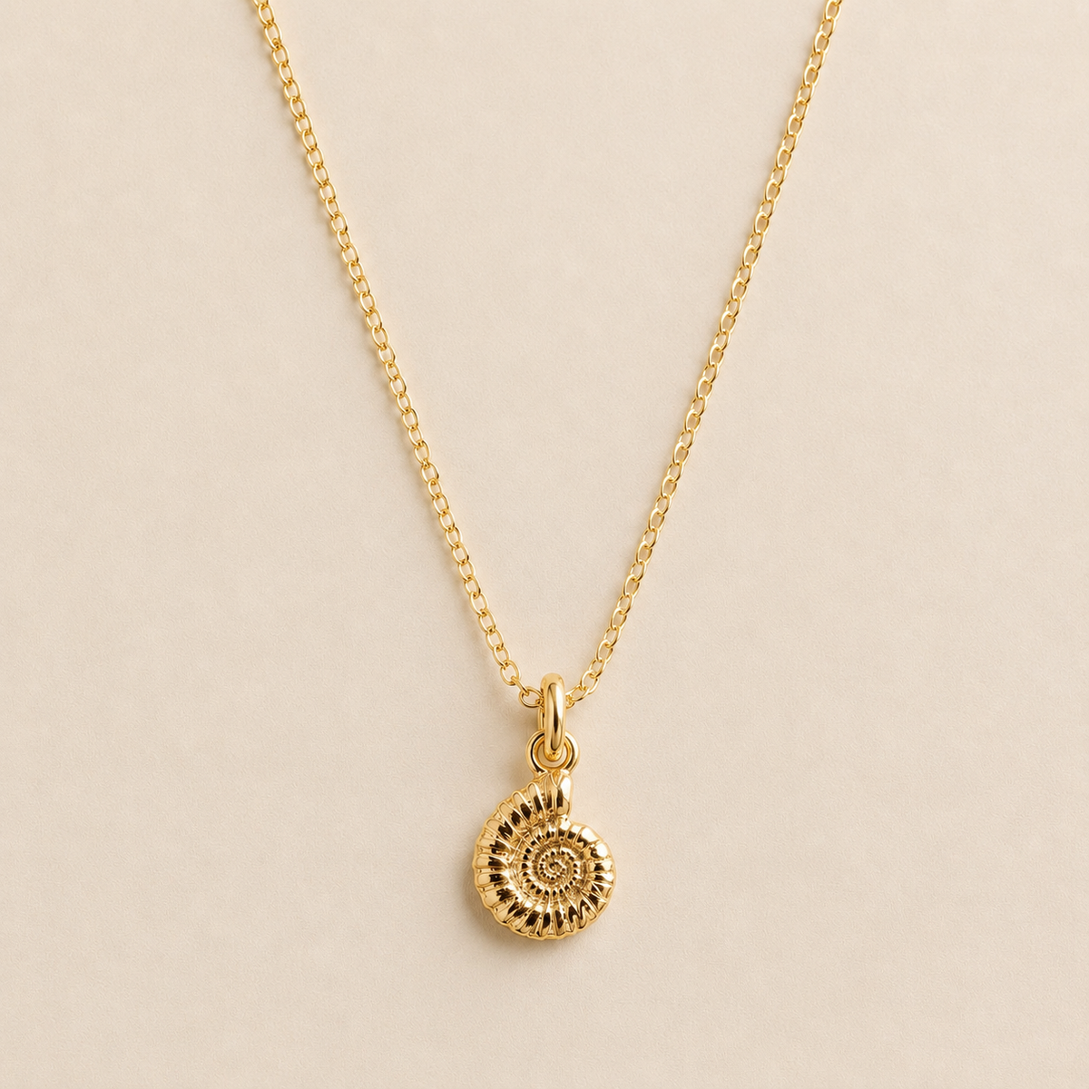 A delicate gold chain necklace featuring a small spiral ammonite fossil pendant, displayed against a solid, warm beige background.