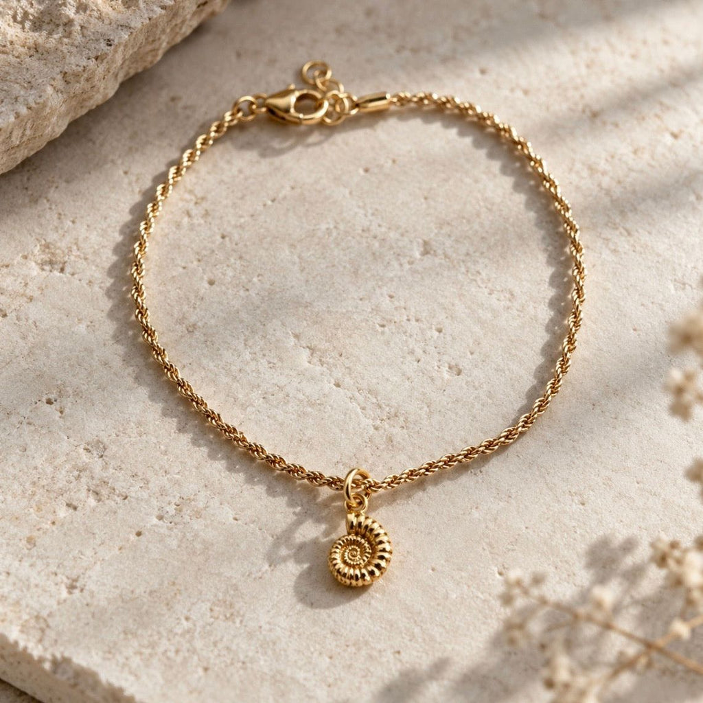 A delicate gold chain bracelet with a small ammonite charm, displayed on a textured beige stone surface with soft shadows.