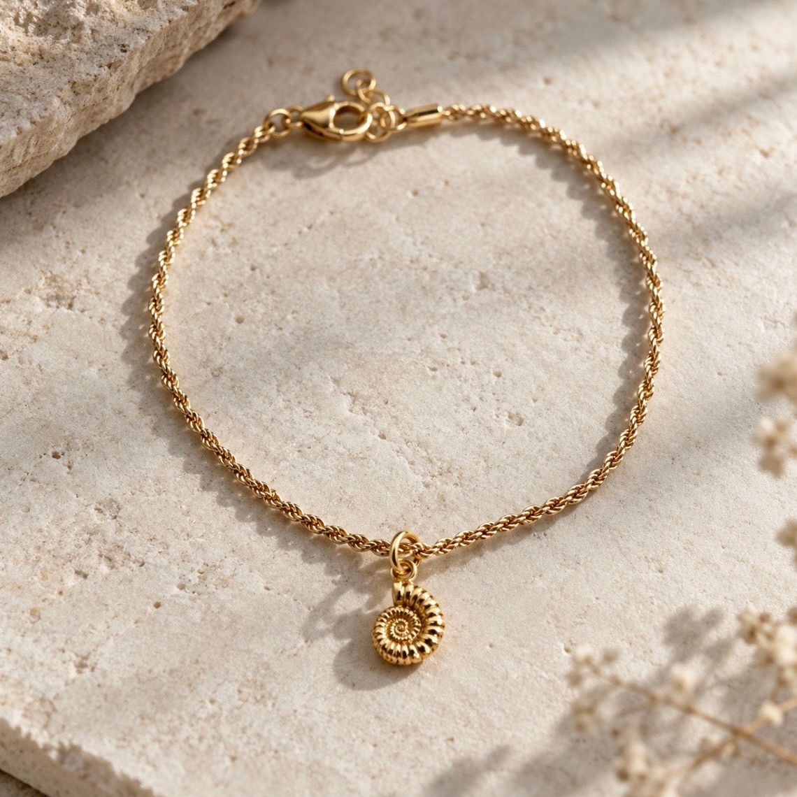 A delicate gold chain bracelet with a small ammonite charm, displayed on a textured beige stone surface with soft shadows.