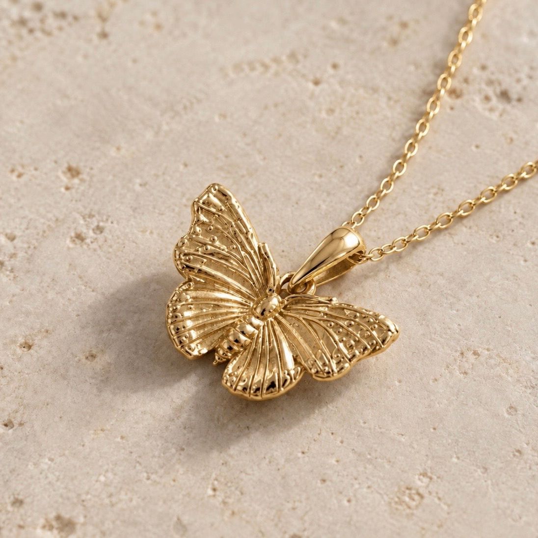 A gold butterfly pendant necklace with a textured wing design, resting on a light beige stone surface.