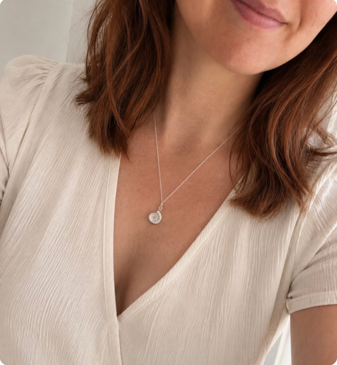A close-up of a woman wearing a delicate silver ammonite fossil  pendant, paired with a cream-colored crinkled blouse.