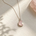 A delicate silver chain necklace with a crescent moon pendant made of light pink rose quartz stone, displayed on a textured cream surface with soft shadows.