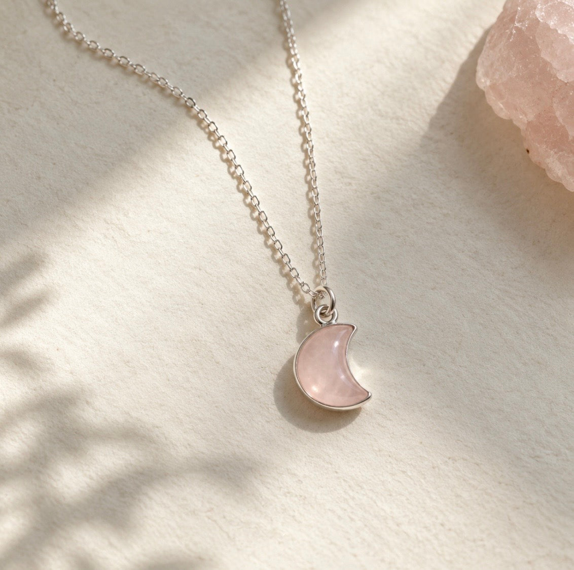 A delicate silver chain necklace with a crescent moon pendant made of light pink rose quartz stone, displayed on a textured cream surface with soft shadows.
