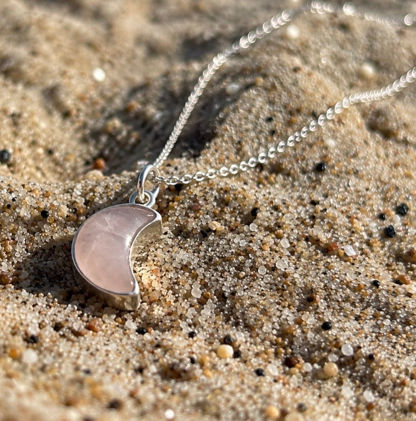 A silver necklace with a crescent moon pendant made of pale pink rose quartz gemstone, resting on sandy ground.