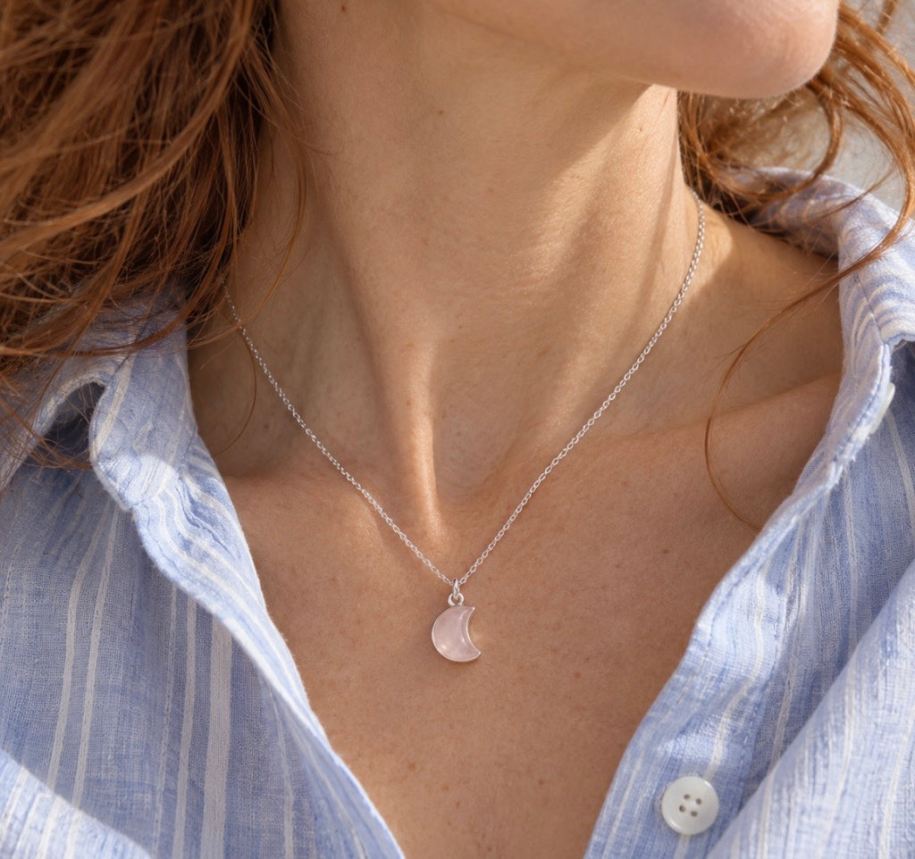 A close-up lifestyle shot of a woman wearing a delicate silver chain necklace with a small crescent moon pendant. The pendant is a pale pinkish rose quartz gemstone  stone, and she is wearing a light blue and white striped button-down shirt.