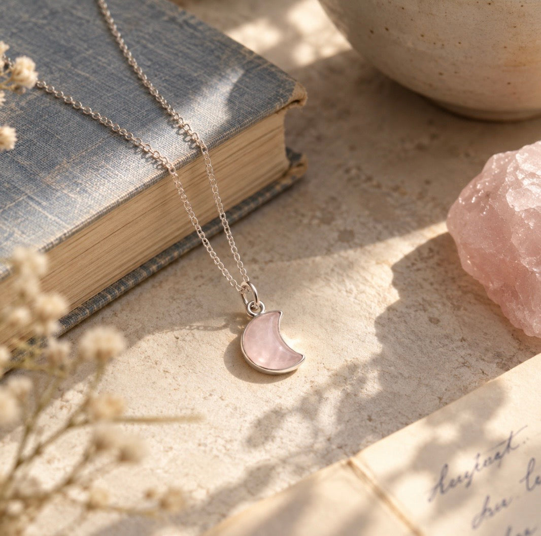 A silver necklace with a crescent moon pendant set in a light pink rose quartz gemstone stone, displayed on a textured beige surface next to a vintage book, dried flowers, and a pink crystal.