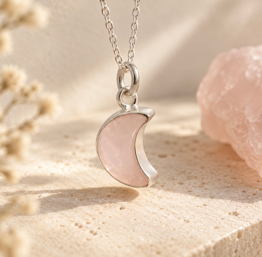 A silver crescent moon pendant necklace with a smooth, polished pale pink rose quartz gemstone , displayed on a textured beige stone surface with a pink crystal and dried flowers in the background.