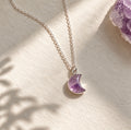 A silver chain necklace with a crescent moon pendant made of purple amethyst, displayed on a textured cream surface with soft shadows and a crystal cluster in the corner.