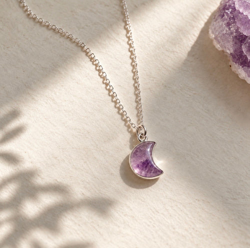 A silver chain necklace with a crescent moon pendant made of purple amethyst, displayed on a textured cream surface with soft shadows and a crystal cluster in the corner.