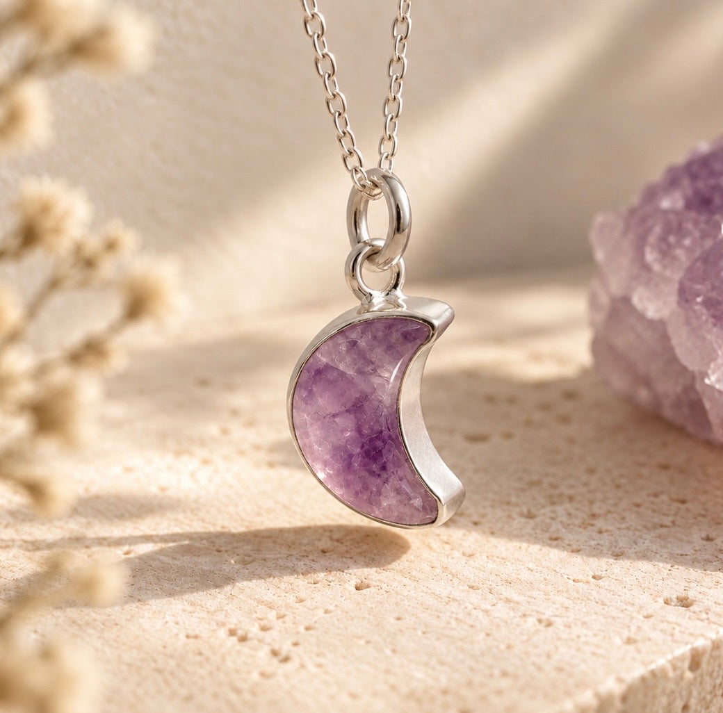 A silver crescent moon pendant necklace with a polished purple amethyst center, resting on a textured beige stone surface with dried flowers and an amethyst crystal in the background.