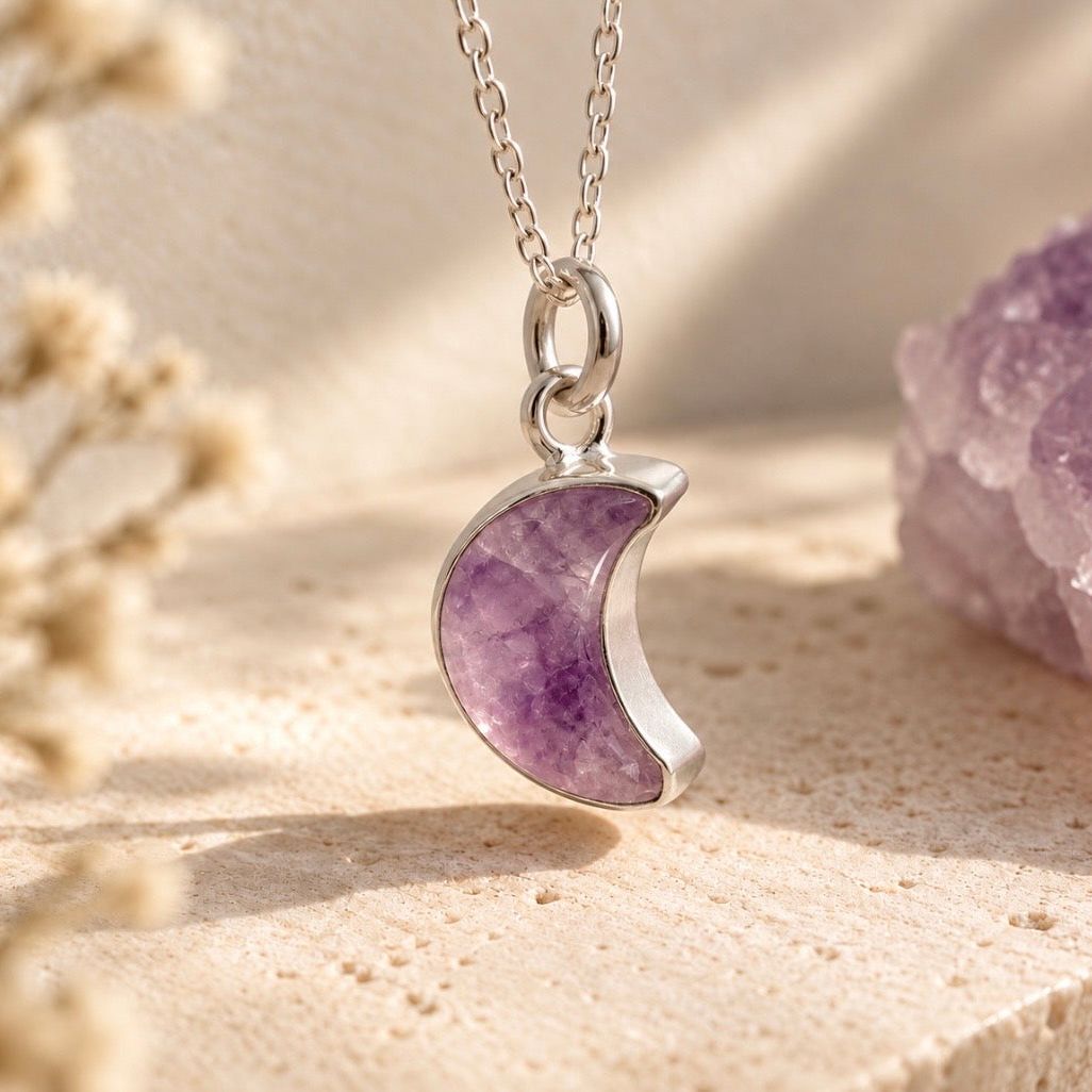 A silver crescent moon pendant necklace with a polished purple amethyst stone, displayed on a textured beige surface with dried flowers and a crystal cluster in the background.