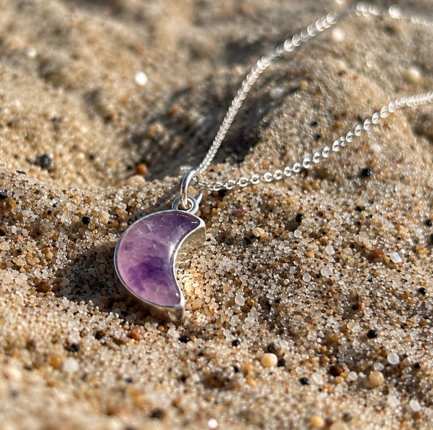 A silver necklace with a crescent moon pendant made of purple amethyst, resting on a sandy beach.