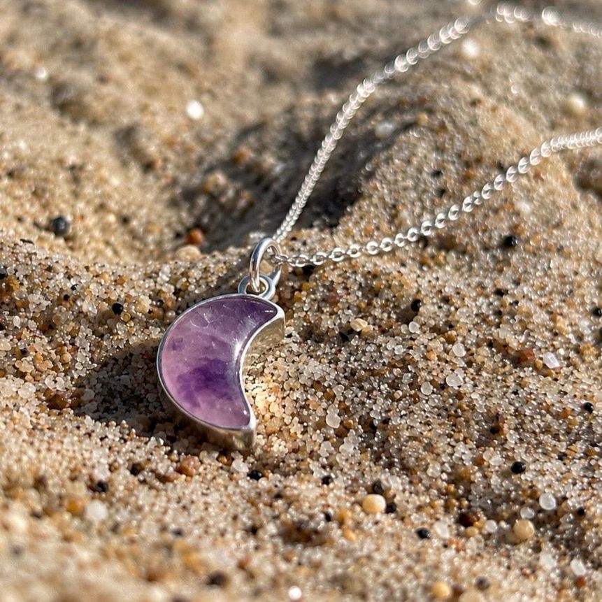 A silver chain necklace with a crescent moon pendant made of purple amethyst, resting on a bed of sand.