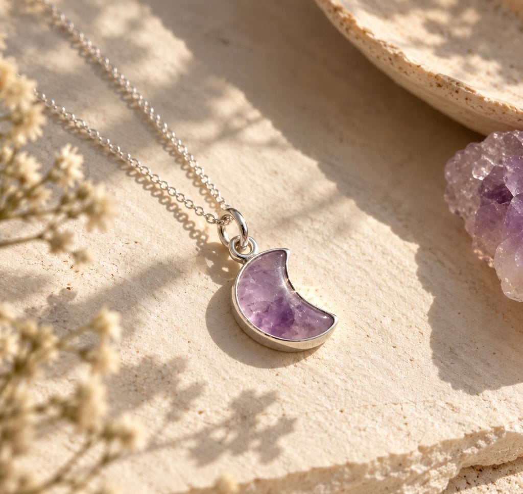 A silver chain necklace with a crescent moon pendant made of purple amethyst, styled on a textured beige stone surface with dried flowers and a crystal cluster.