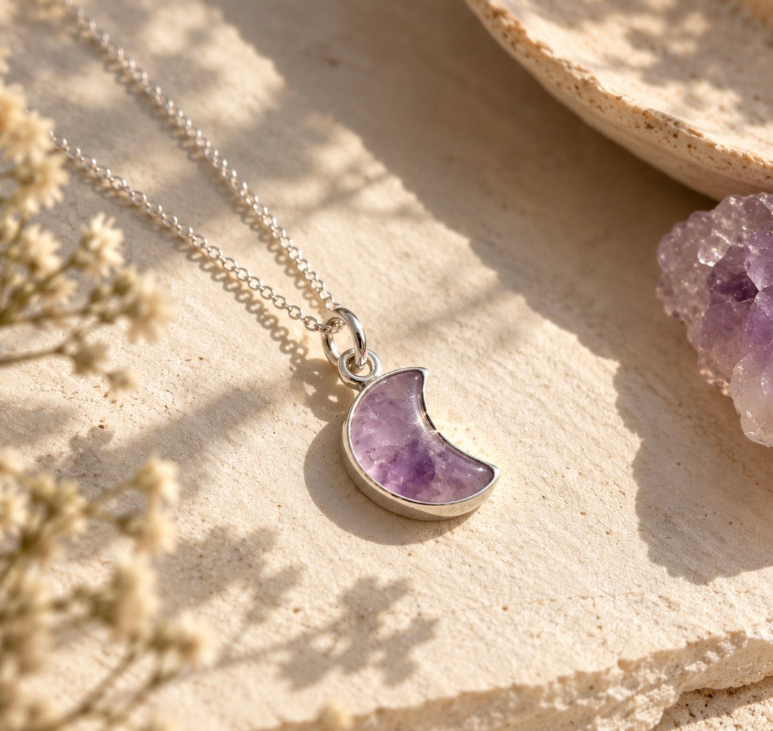 A silver chain necklace with a crescent moon pendant made of purple amethyst, styled on a textured beige stone surface with dried flowers and a crystal cluster.