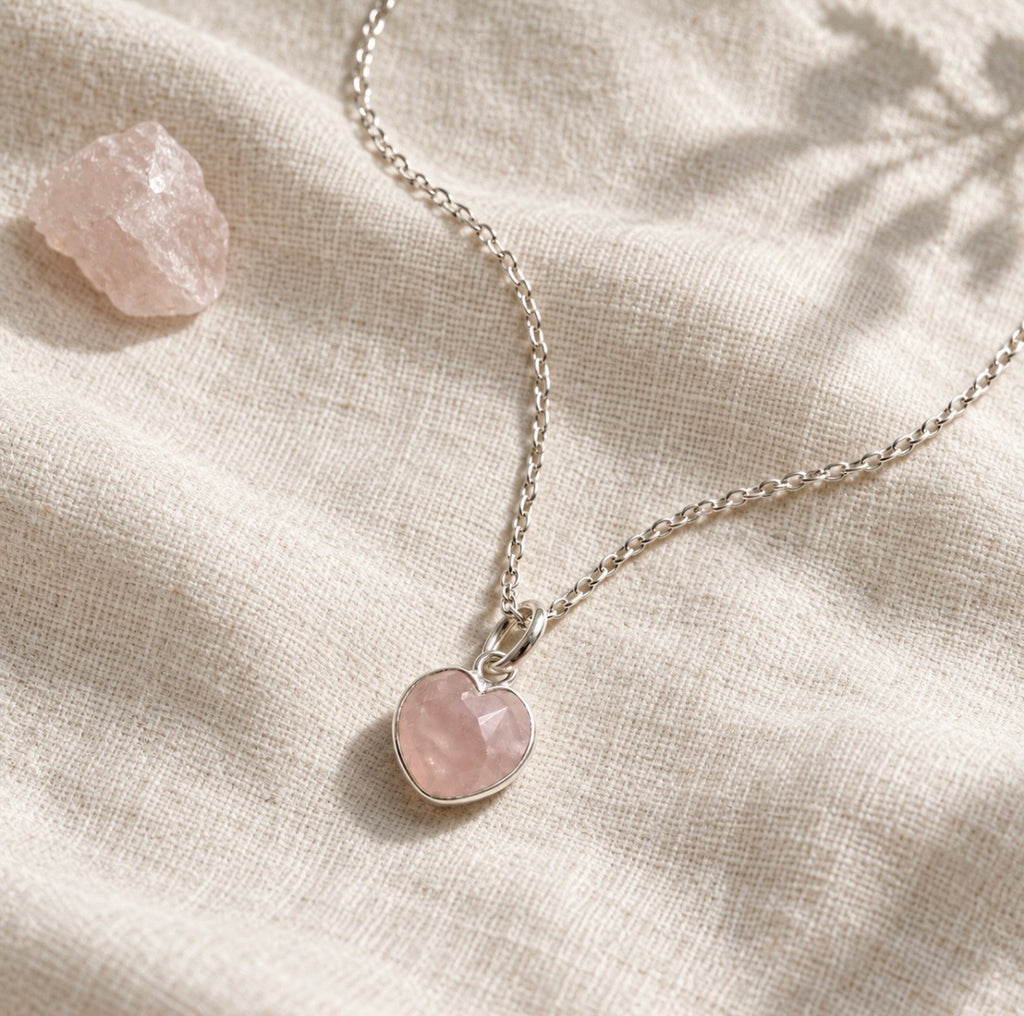 A silver heart-shaped pendant necklace with a faceted light pink rose quartz stone, resting on a textured beige linen fabric next to a raw rose quartz crystal.