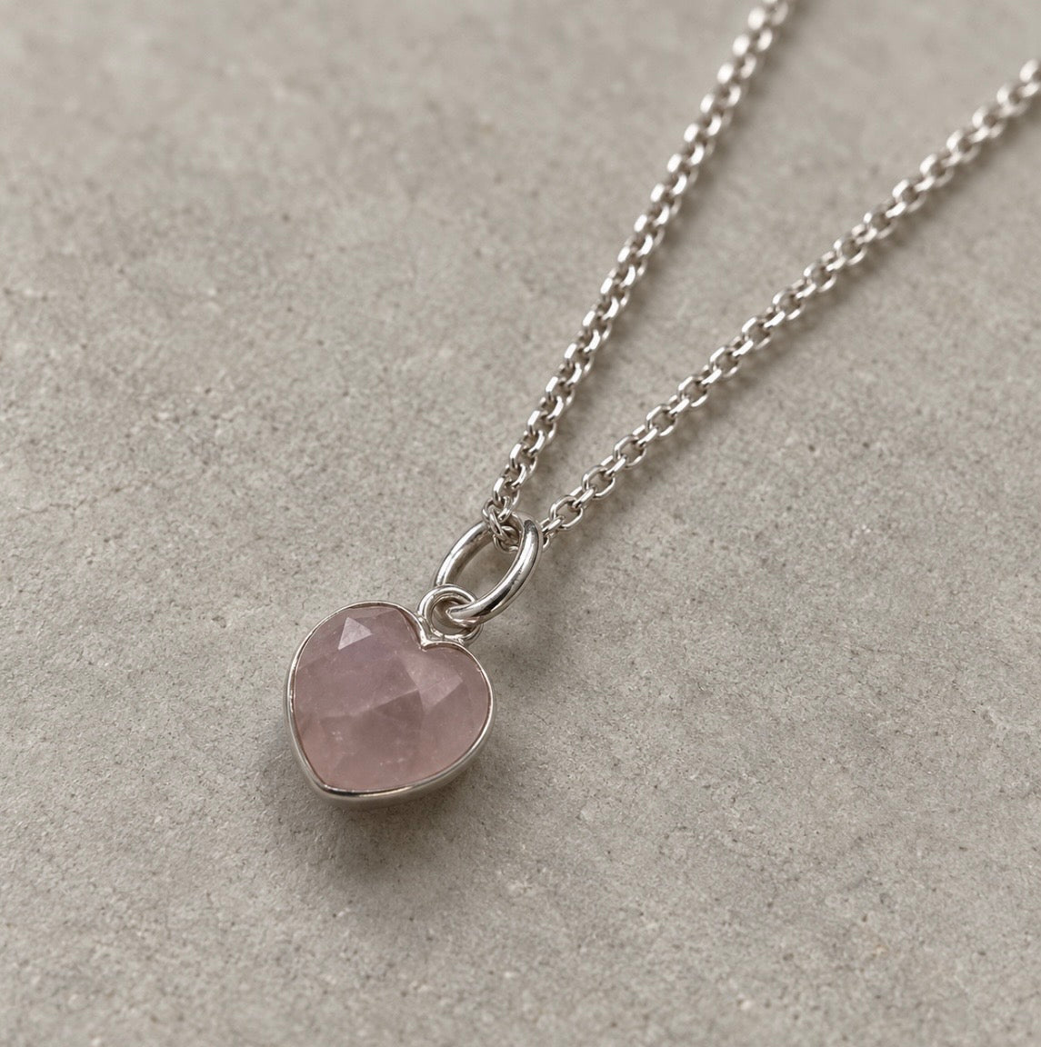 A silver chain necklace with a heart-shaped faceted pink rose quartz gemstone pendant, displayed on a textured gray surface.