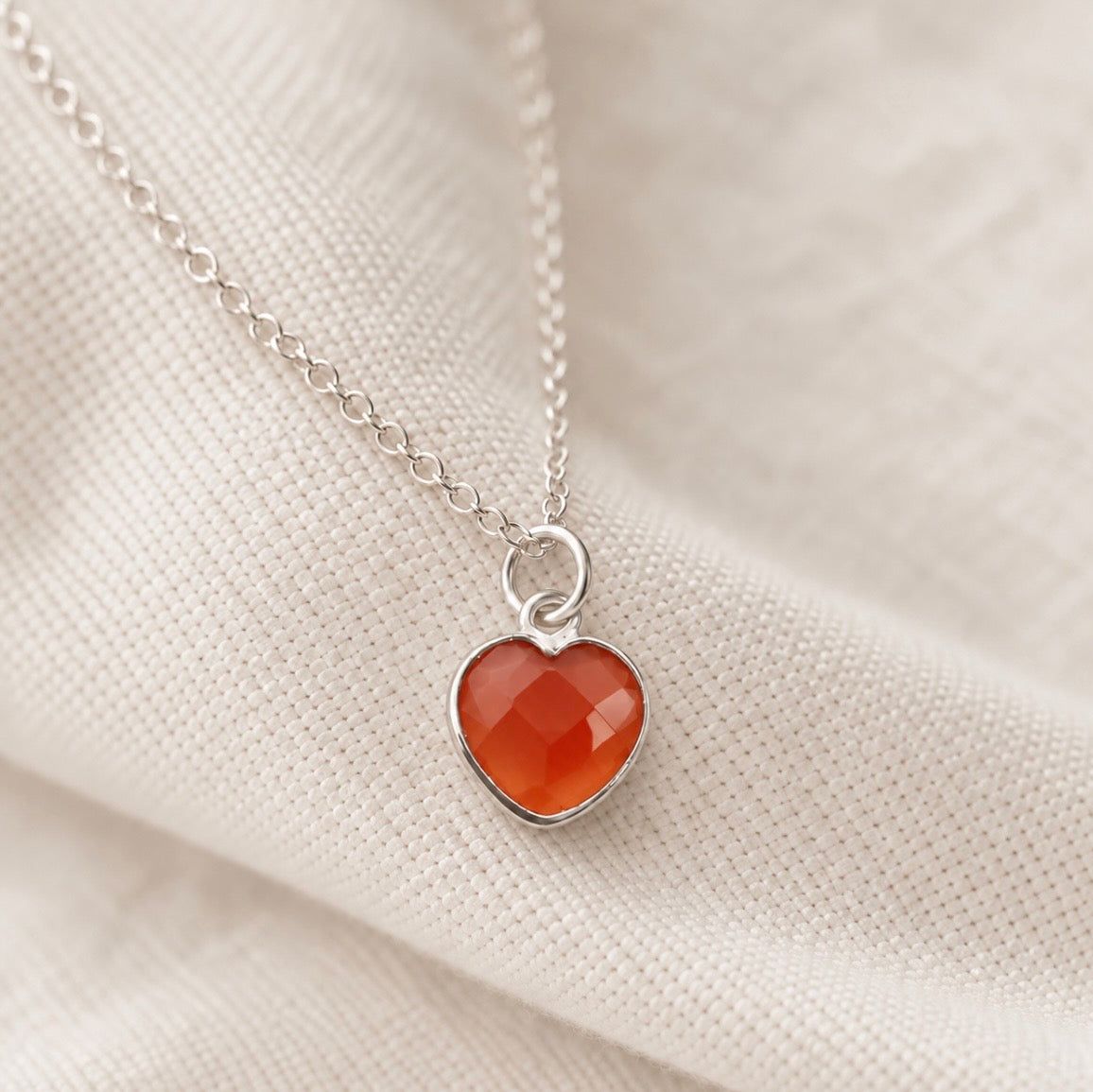 A silver heart-shaped pendant necklace with a faceted orange-red carnelian gemstone, resting on a textured beige fabric background.