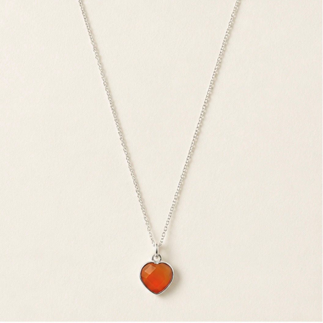 A delicate silver chain necklace featuring a small heart-shaped pendant filled with a vibrant orange-red carnelian stone, set against a plain off-white background.