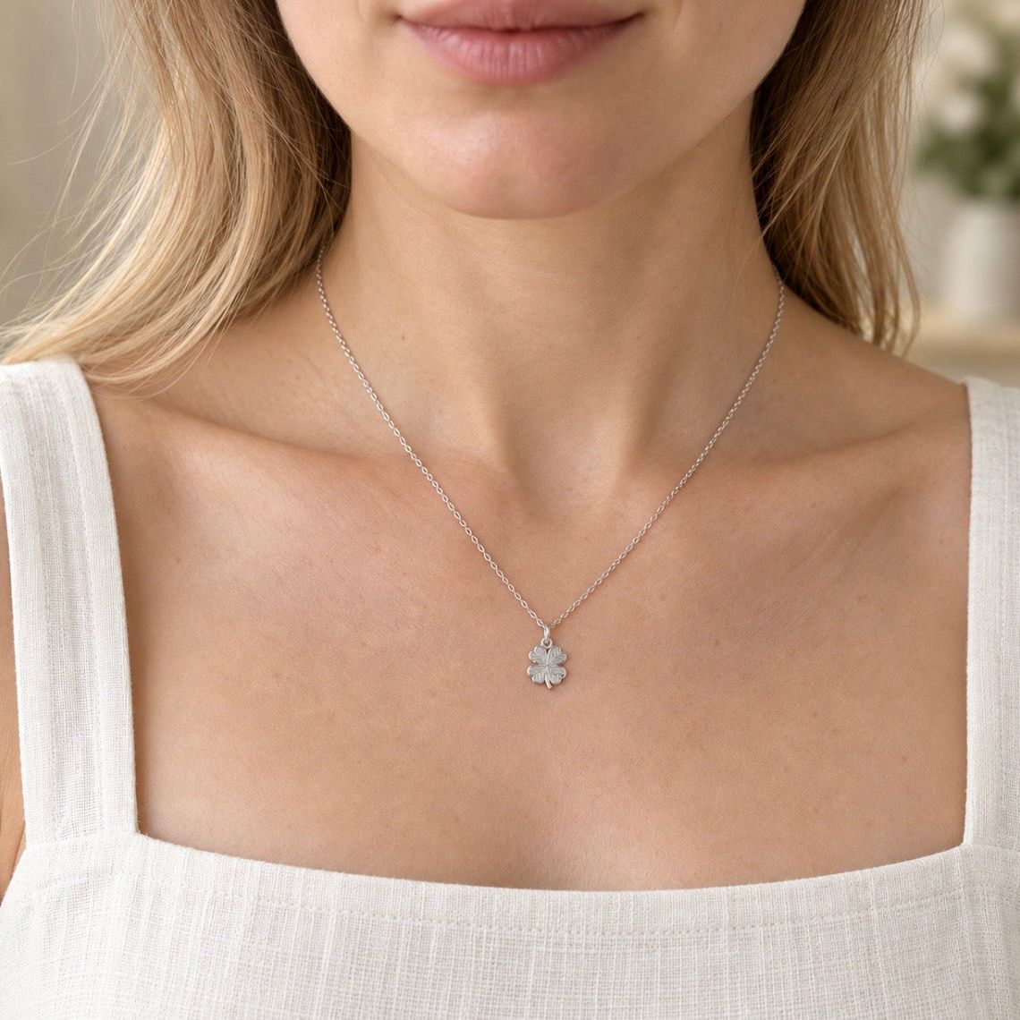 A close-up of a woman wearing a delicate silver chain necklace with a small silver clover pendant, styled with a white linen top.