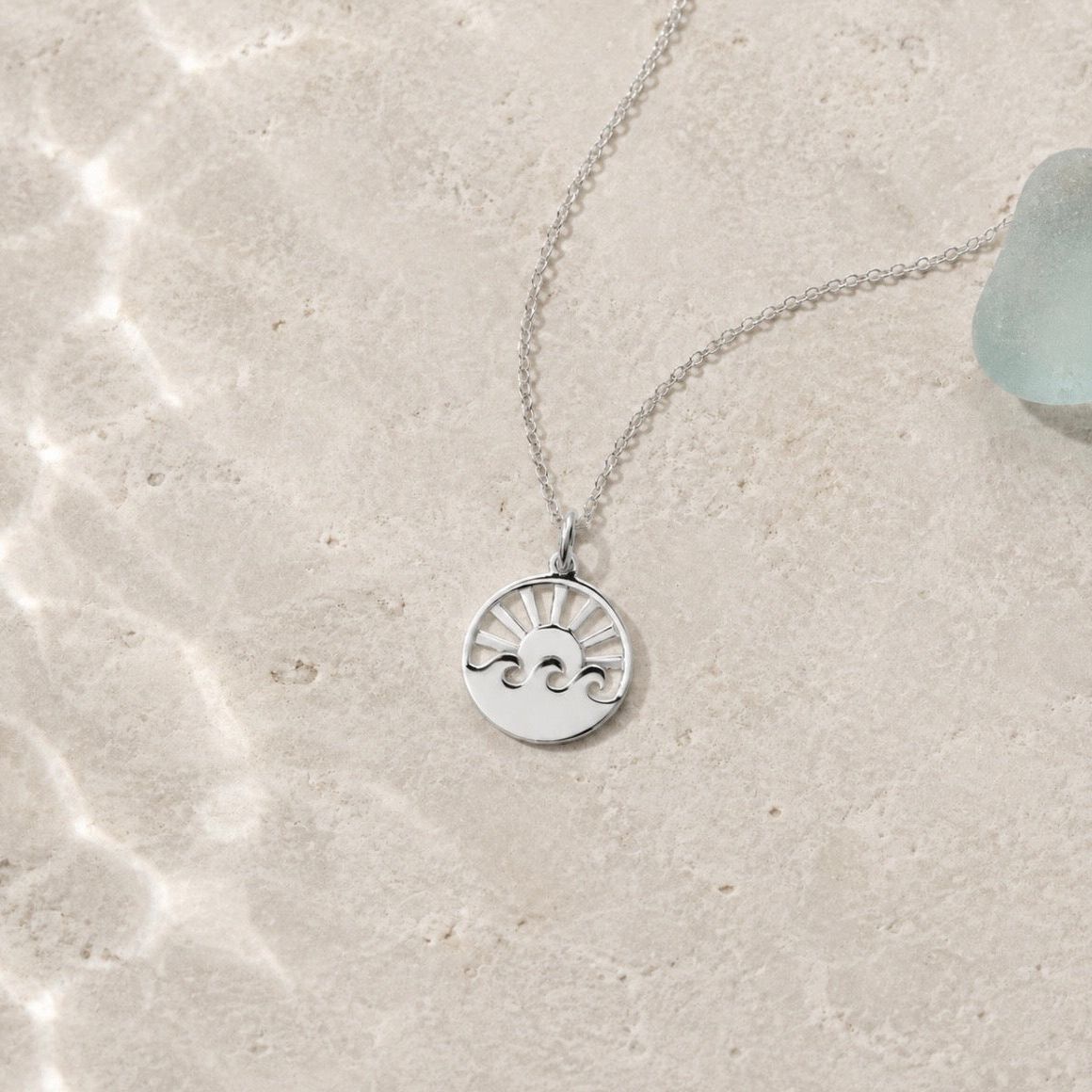 A silver pendant necklace featuring a sunburst and wave design, displayed on a textured beige stone surface with soft natural light and a piece of sea glass in the corner.