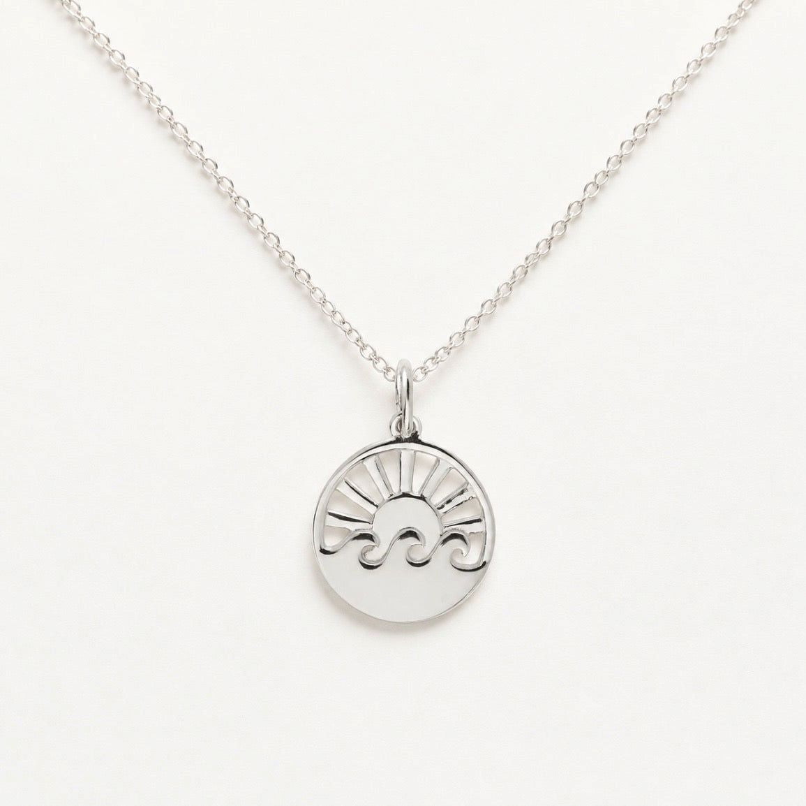 A silver pendant necklace featuring a circular disc charm with a stylized sun and wave design, suspended from a delicate silver chain against a clean white background.