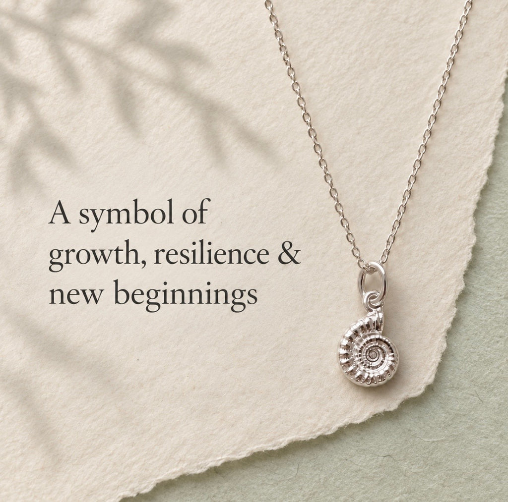 A silver necklace with a spiral ammonite pendant is displayed on a textured cream paper background with a torn-edge overlay and soft leaf shadows. Text on the left reads, 'A symbol of growth, resilience & new beginnings'.
