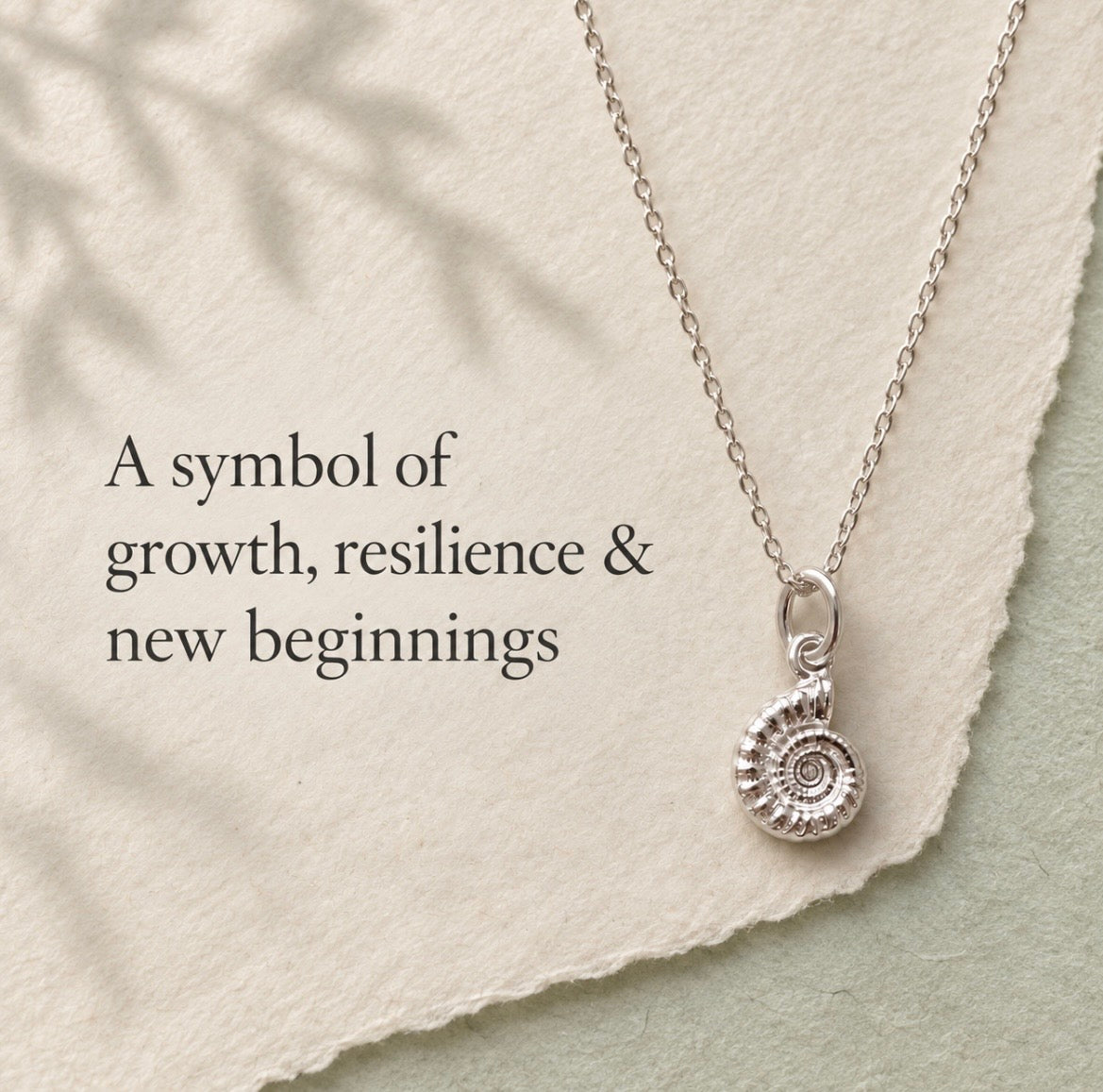 A silver necklace with a spiral ammonite pendant is displayed on a textured cream paper background with a torn-edge overlay and soft leaf shadows. Text on the left reads, 'A symbol of growth, resilience & new beginnings'.