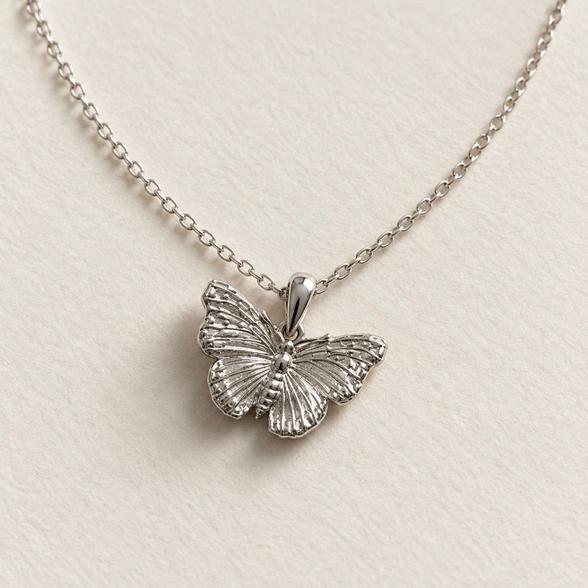 A silver butterfly pendant necklace on a delicate silver chain, displayed on a textured off-white surface.