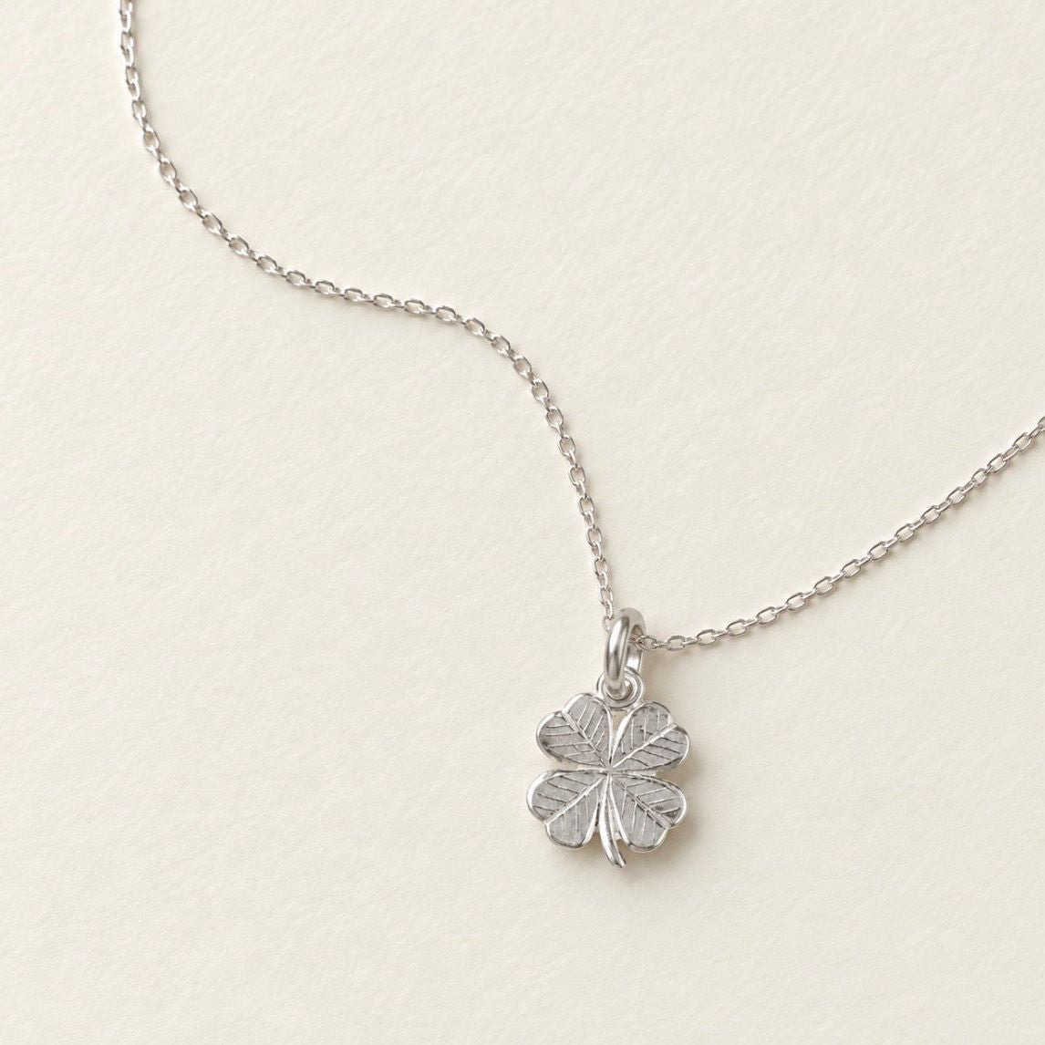 A sterling silver four-leaf clover pendant necklace on a delicate cable chain, displayed on a textured off-white background.