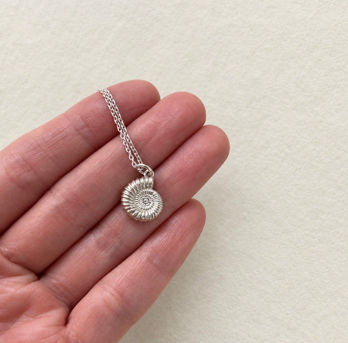 A silver ammolite fossil pendant necklace held on a person's fingers against a light beige textured background.