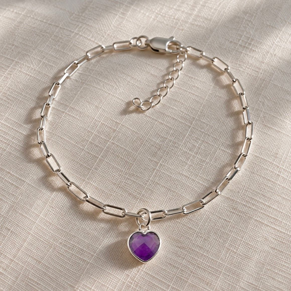 A silver paperclip chain bracelet with a heart-shaped purple amethyst gemstone pendant, displayed on a textured beige fabric background.