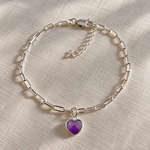 A silver paperclip chain bracelet with a heart-shaped purple amethyst gemstone pendant, displayed on a textured beige fabric background.