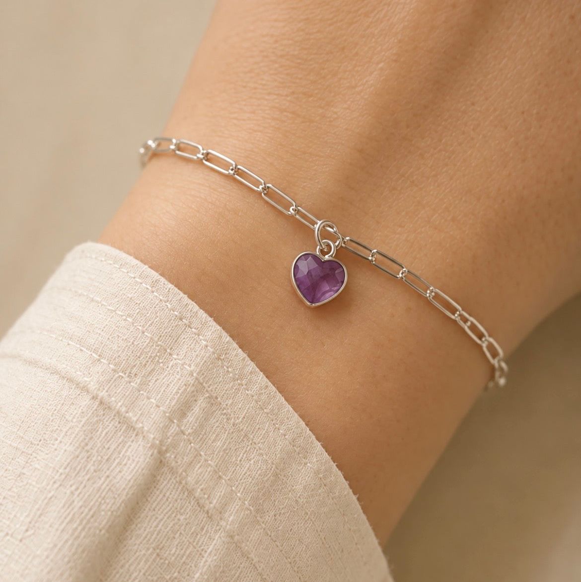 A delicate silver chain bracelet with a small heart-shaped purple amethyst gemstone charm, shown worn on a person's wrist.