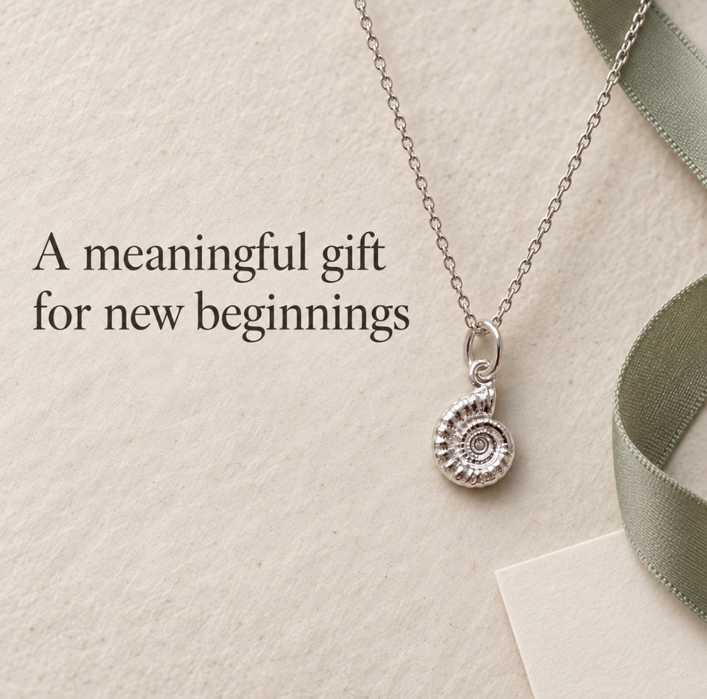 A silver pendant necklace with a spiral ammonite fossil charm, styled on a textured beige background with a green ribbon and the text 'A meaningful gift for new beginnings'.