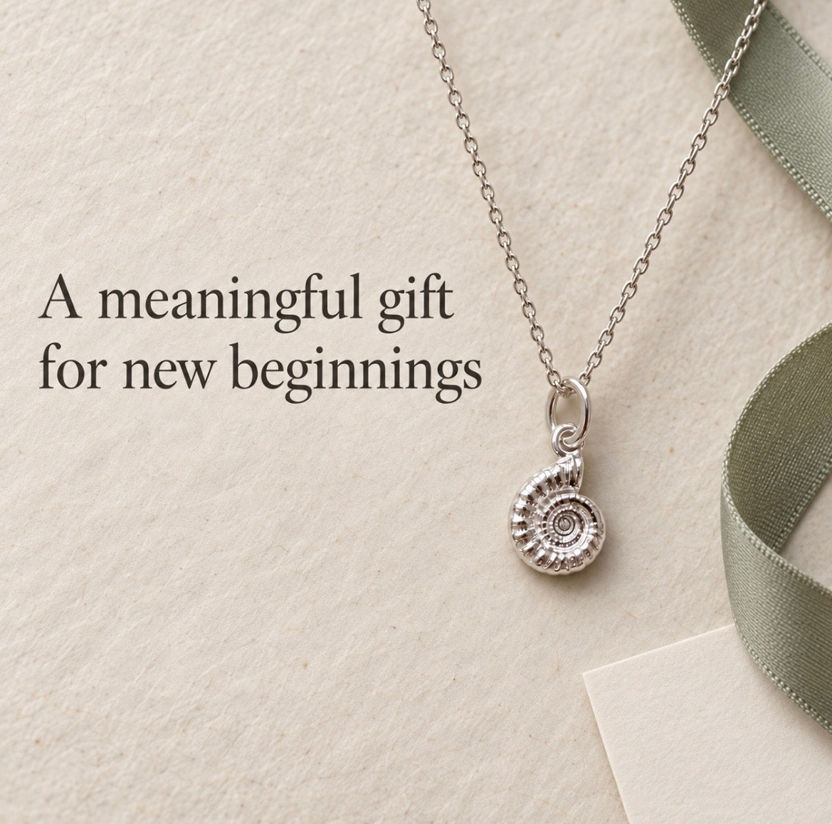 A silver pendant necklace with a spiral ammonite fossil charm, styled on a textured beige background with a green ribbon and the text 'A meaningful gift for new beginnings'.